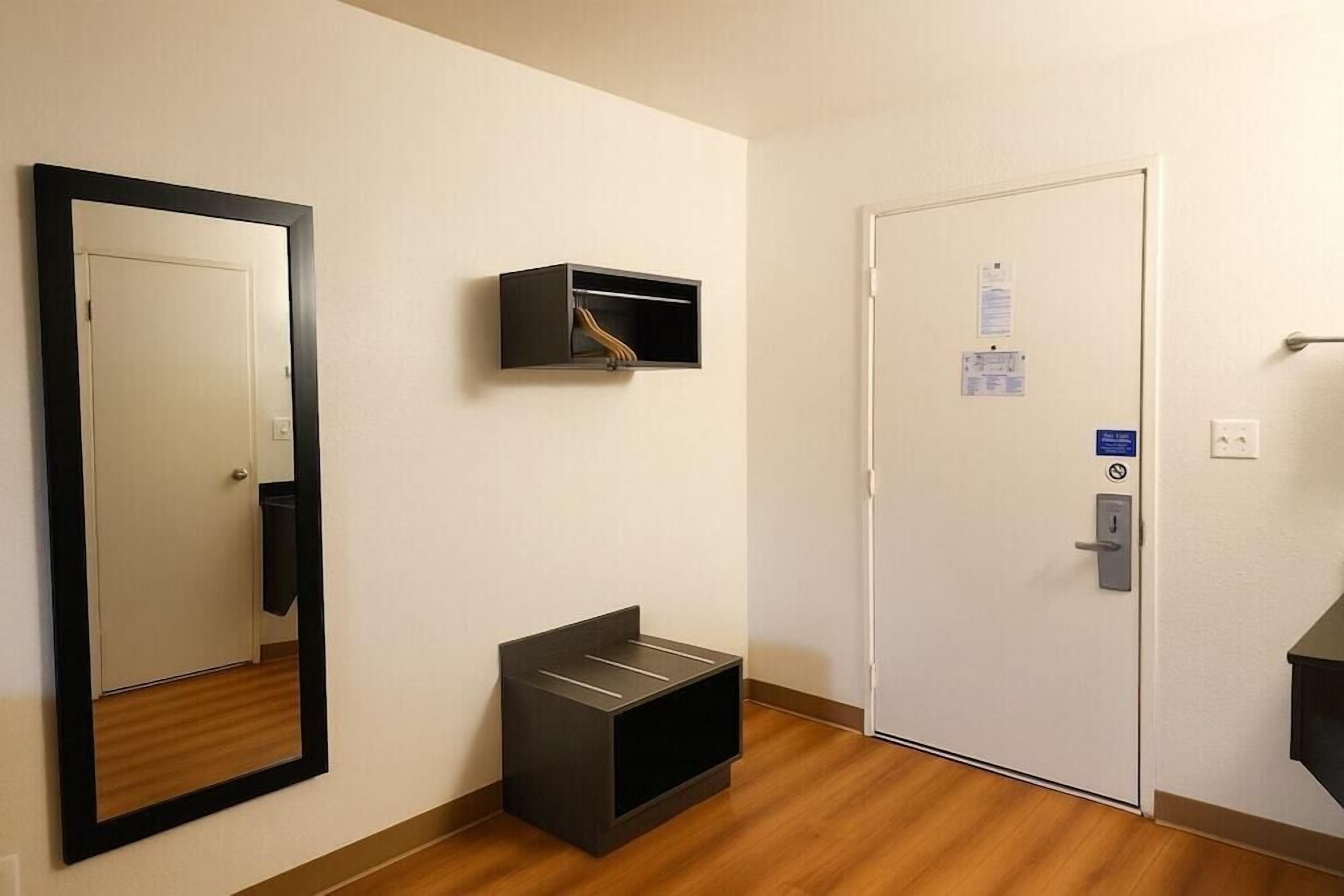 A hotel room with a wooden floor, a large mirror, a black storage unit, and a door with a control panel.