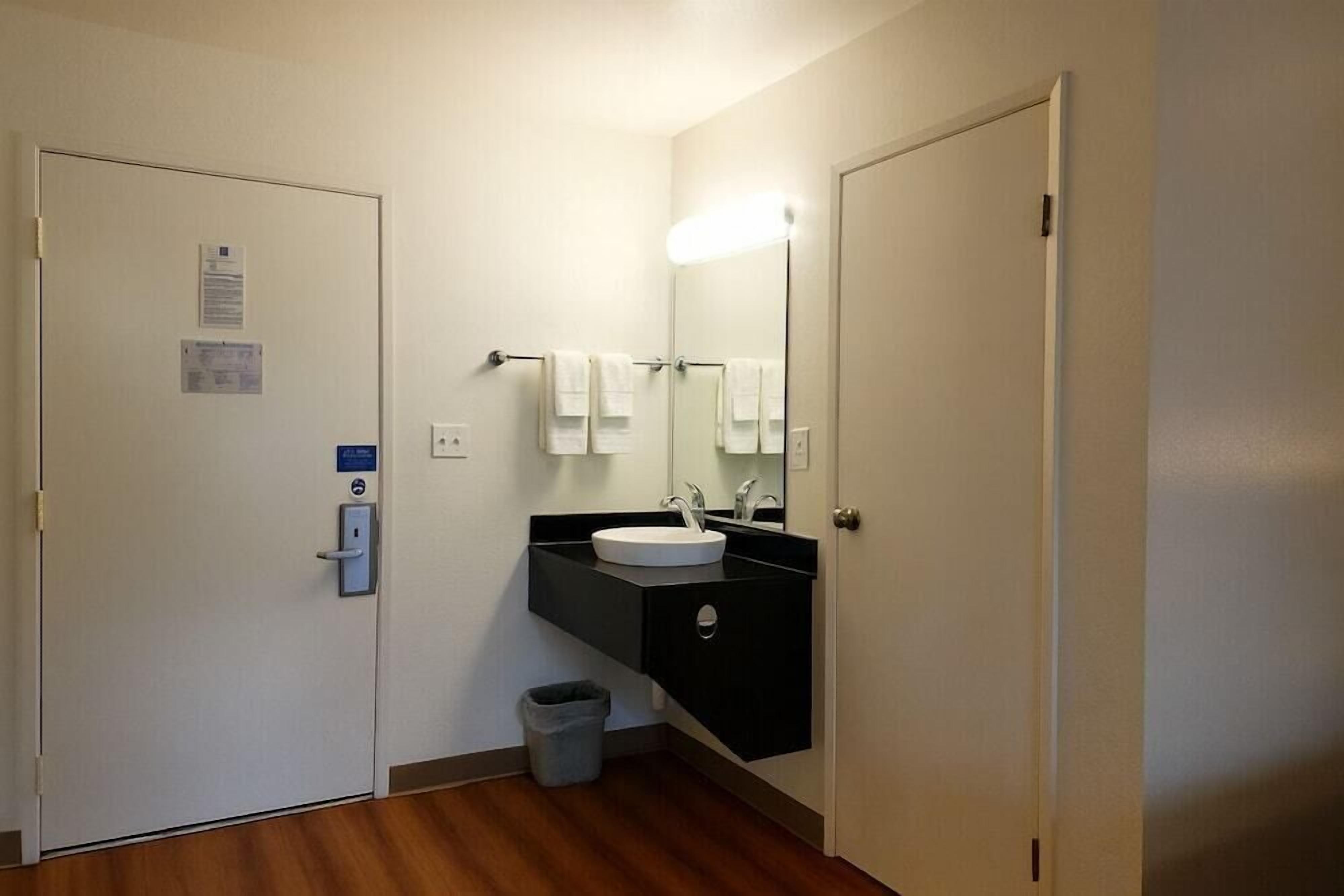 Un baño pequeño con un lavamanos blanco, encimera negra y un espejo encima. Hay un cubo de basura debajo del lavamanos y una puerta con cerradura a la izquierda.