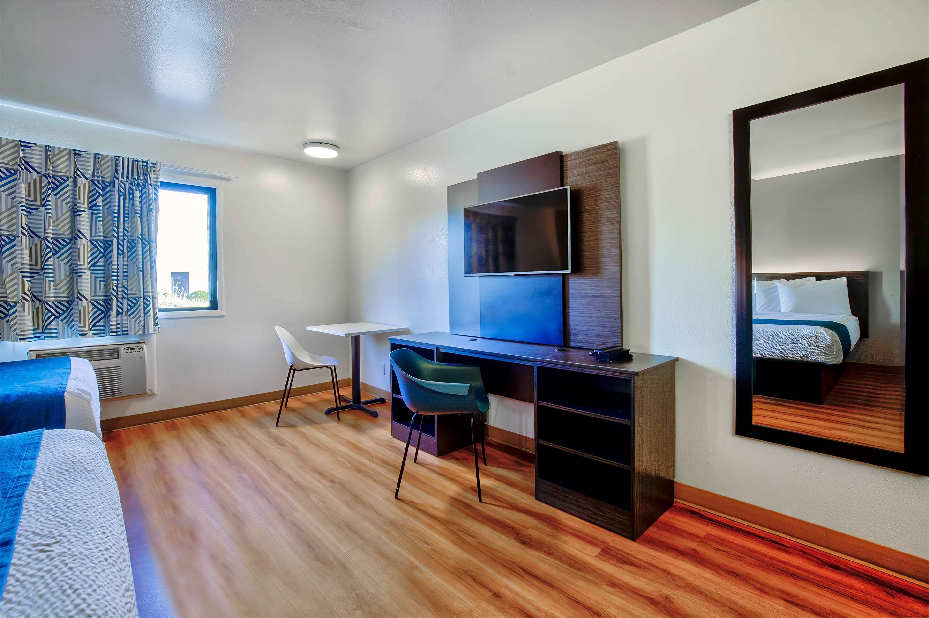 A modern hotel room with a wooden floor, a desk with a chair, a bed, a large mirror, and a TV mounted on the wall.