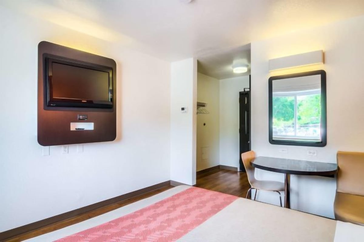 Ett hotellrum med en säng, ett skrivbord med en stol, en tv som är monterad på väggen och ett fönster.
