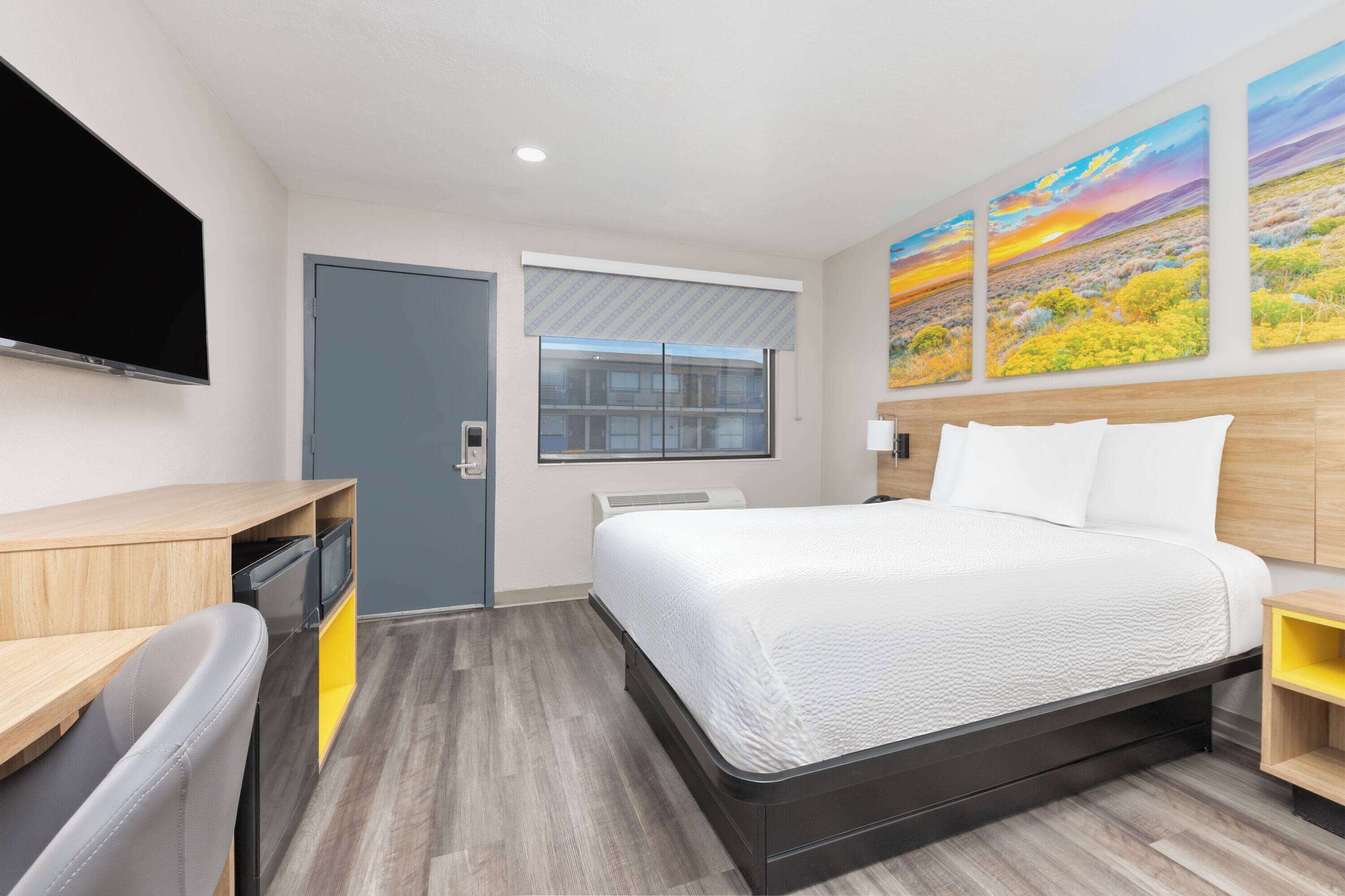 A modern hotel room with a bed, a desk, a chair, a TV, and a window with blinds.