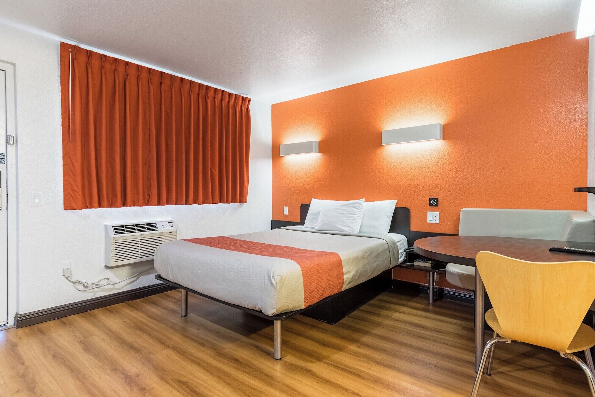Habitación de hotel con cama, escritorio y silla. Se observa un equipo de aire acondicionado en la pared y una ventana con cortinas.