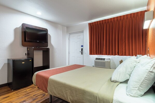 Ein Hotelzimmer mit einem Bett, einem an der Wand befestigten Fernseher, einer kleinen Kühlbox und einem Fenster mit Vorhängen.