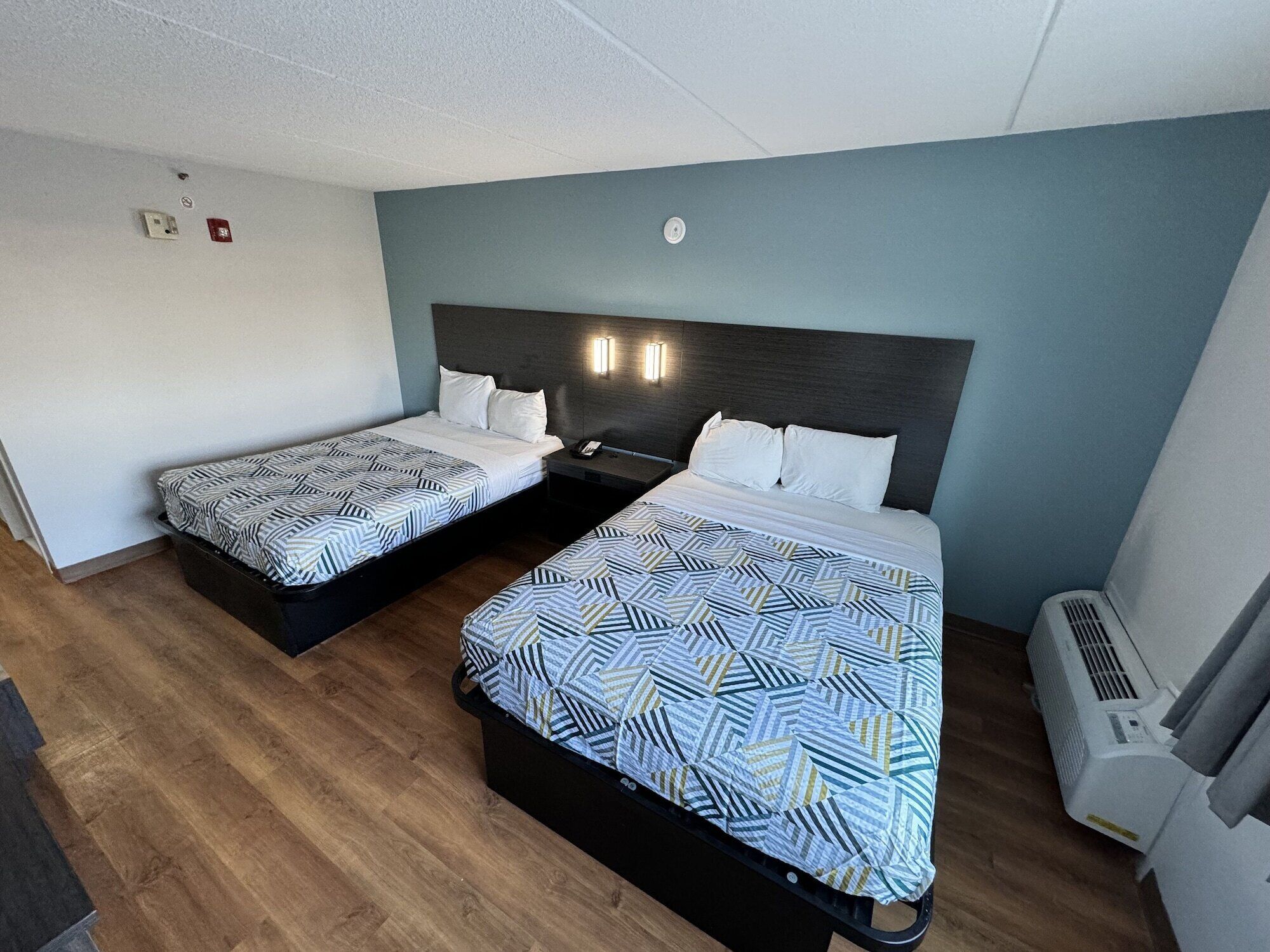 A hotel room with two beds, a wooden floor, a wall-mounted heater, and a light switch.