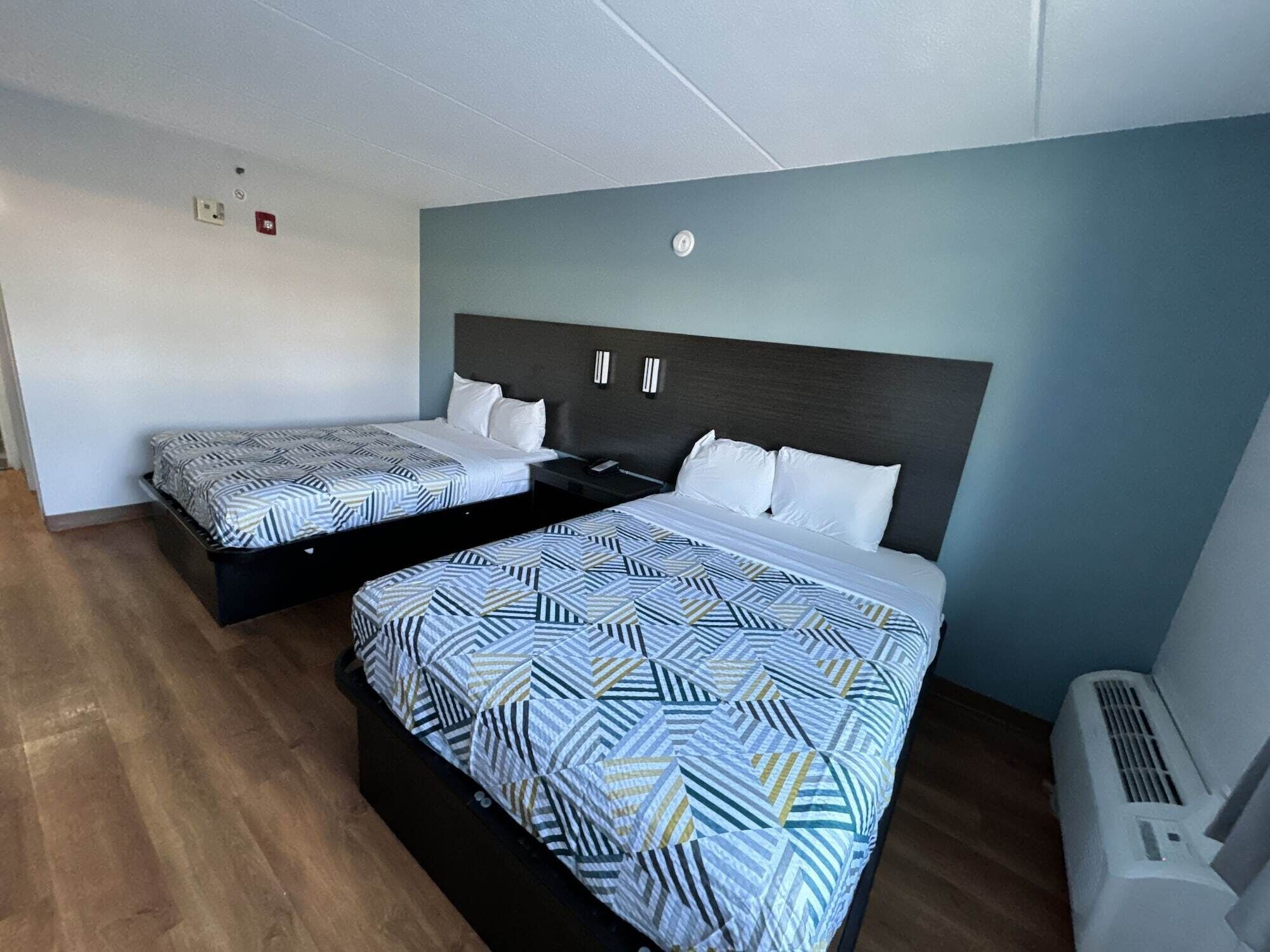 Una habitación de hotel con dos camas, piso de madera, aire acondicionado de pared y una mesita entre las camas.
