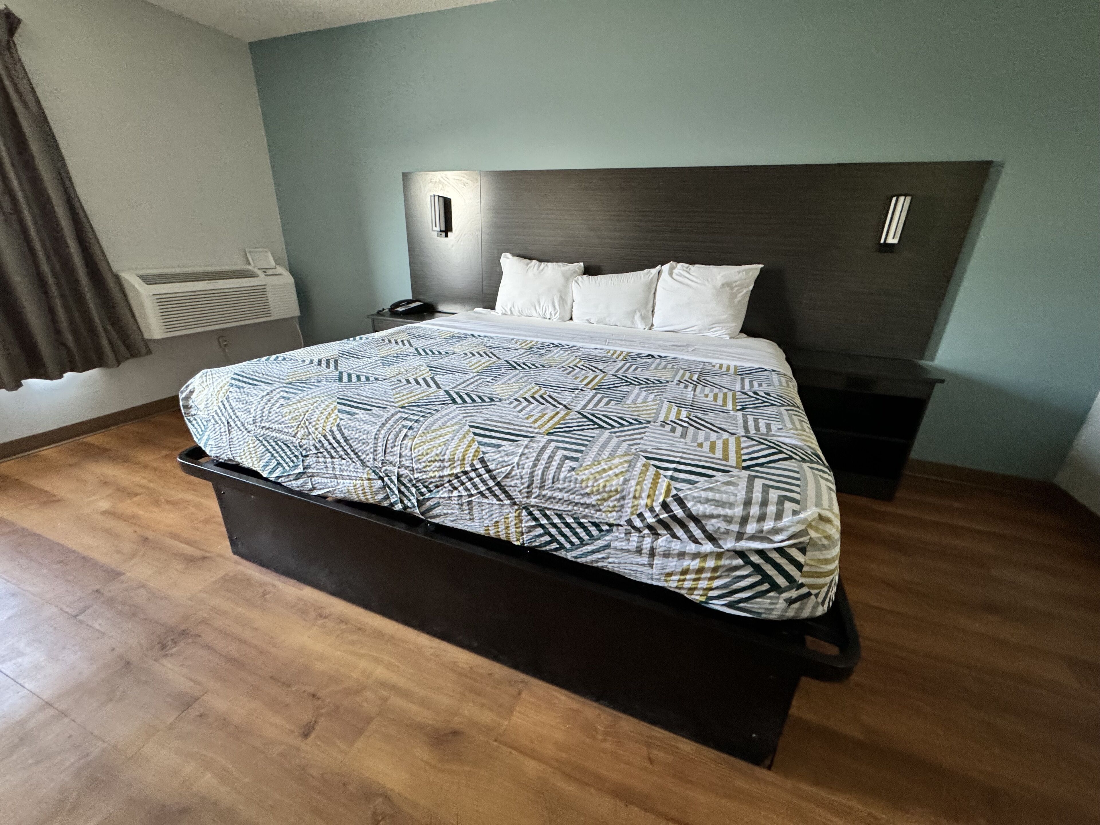 A hotel room with a bed, a nightstand, a wall-mounted air conditioner, and a window with curtains.