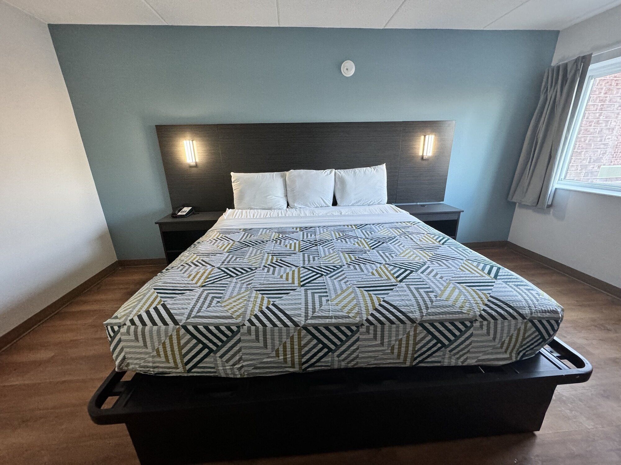 A modern hotel room with a large bed, a headboard with integrated lighting, a bedside table, and a window with curtains.