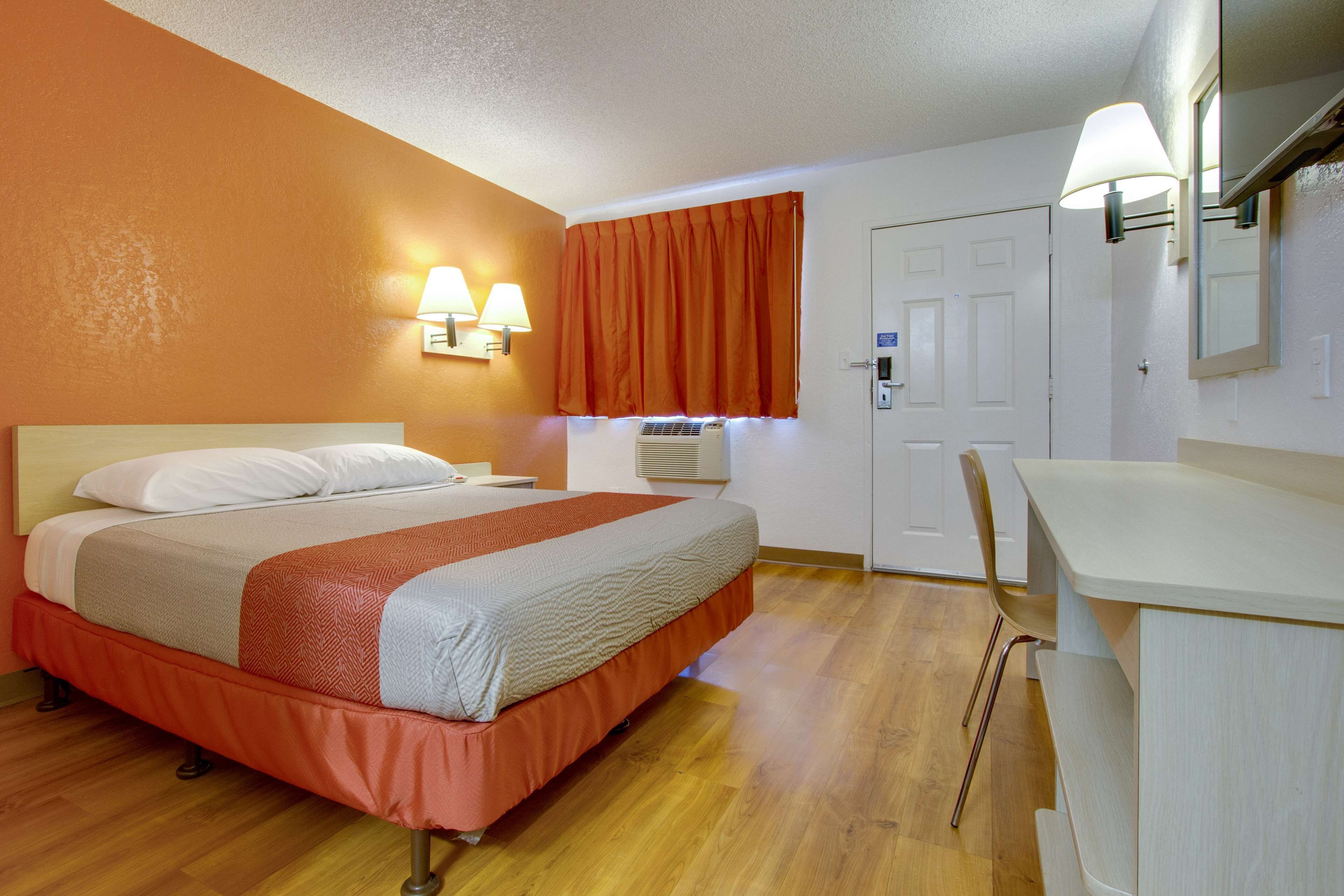 Ett hotellrum med en säng, ett skrivbord, en stol, ett fönster med gardiner och väggmonterade lampor.