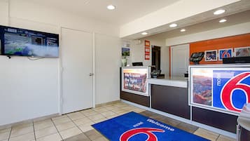 Zona de recepción de hotel con pantalla digital que muestra información meteorológica, mostrador de recepción, felpudo azul y señalización en la pared.