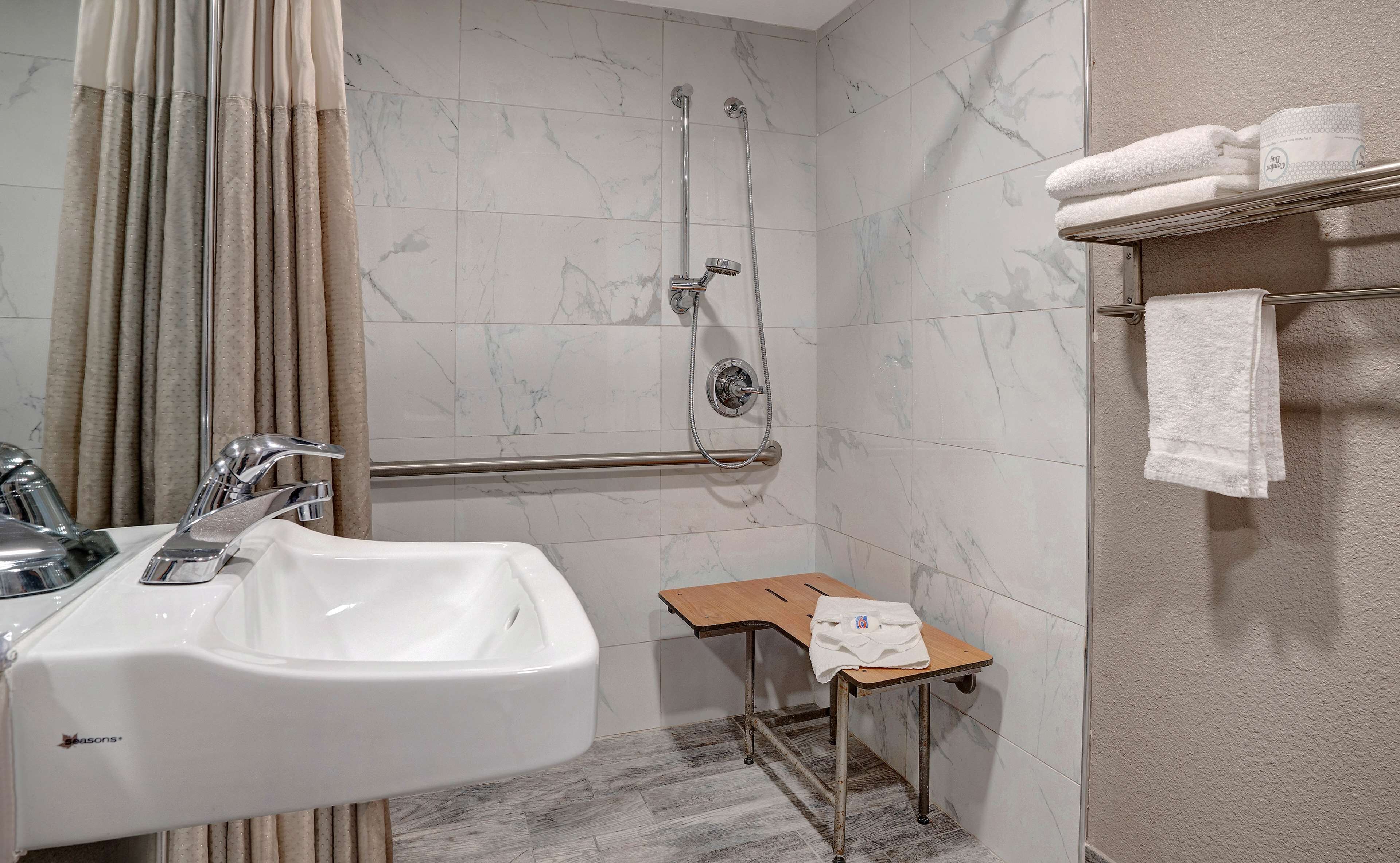 A bathroom with a white sink, a shower with a handheld showerhead, a grab bar, and a small wooden stool with a towel.