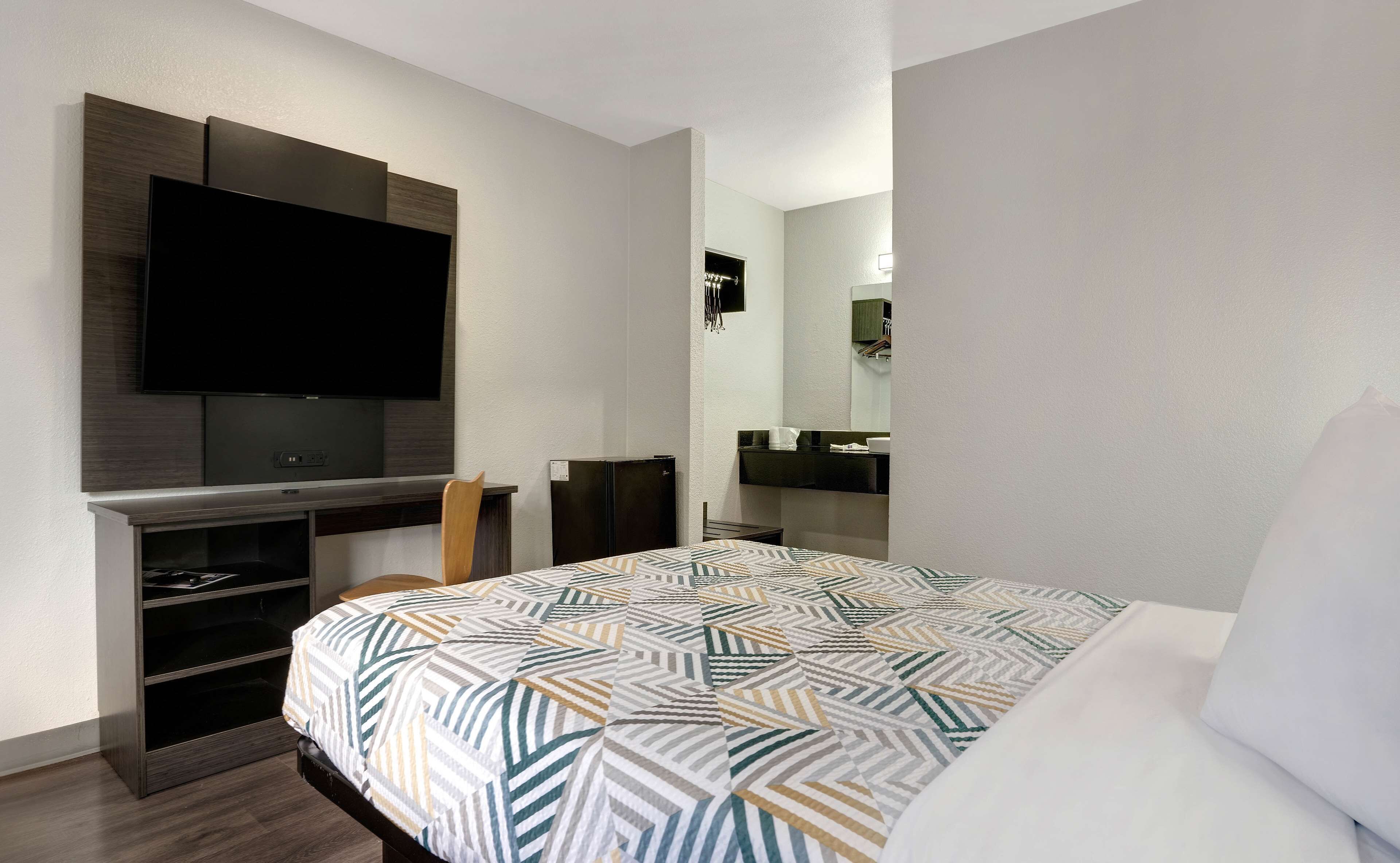 A hotel room with a bed, a TV mounted on the wall, a small desk with a chair, and a mirror above the mirror.
