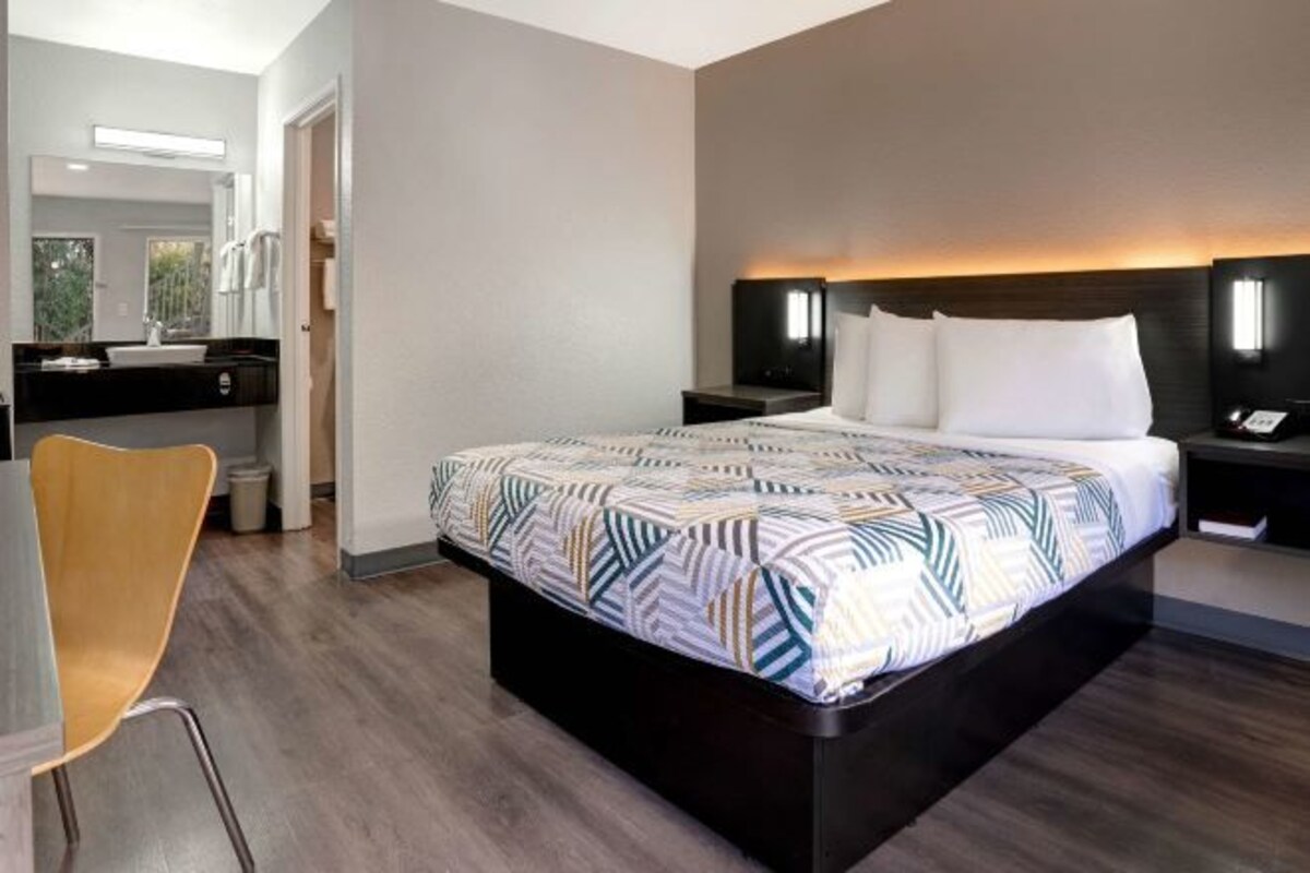A modern hotel room with a large bed, a bedside table, a chair, and a bathroom visible through an open door.
