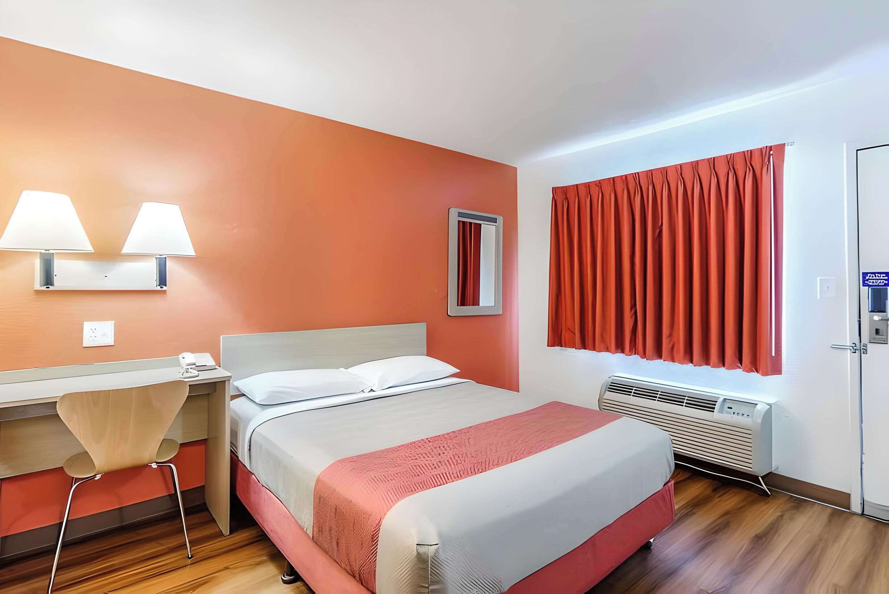 A hotel room with a bed, desk, chair, wall-mounted lamps, a window with curtains, and an air conditioning unit.