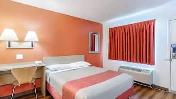 A hotel room with a bed, desk, chair, wall-mounted lamps, a window with curtains, and an air conditioning unit.