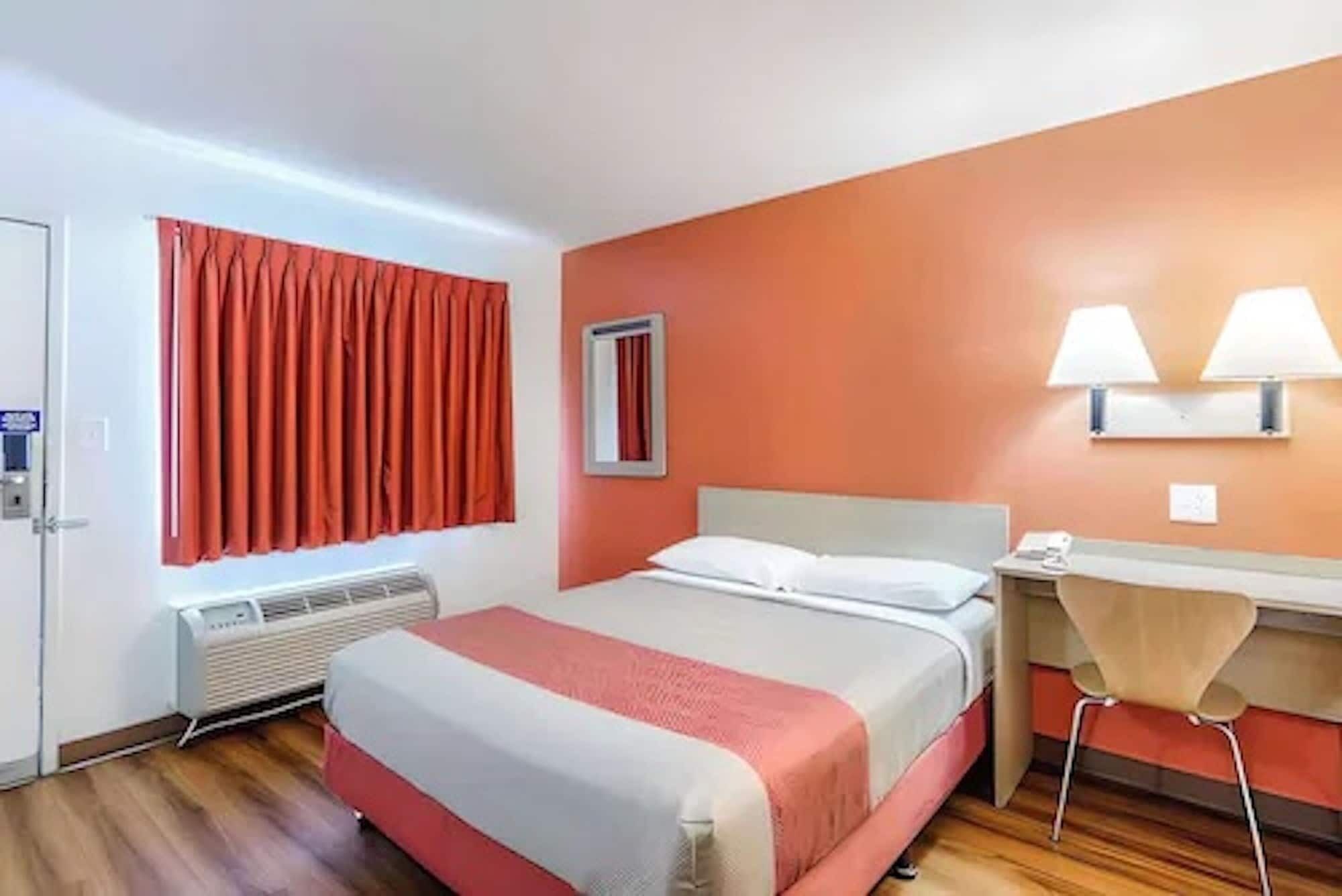 A hotel room with a bed, a desk, a chair, a window with curtains, and a wall-mounted lamp.