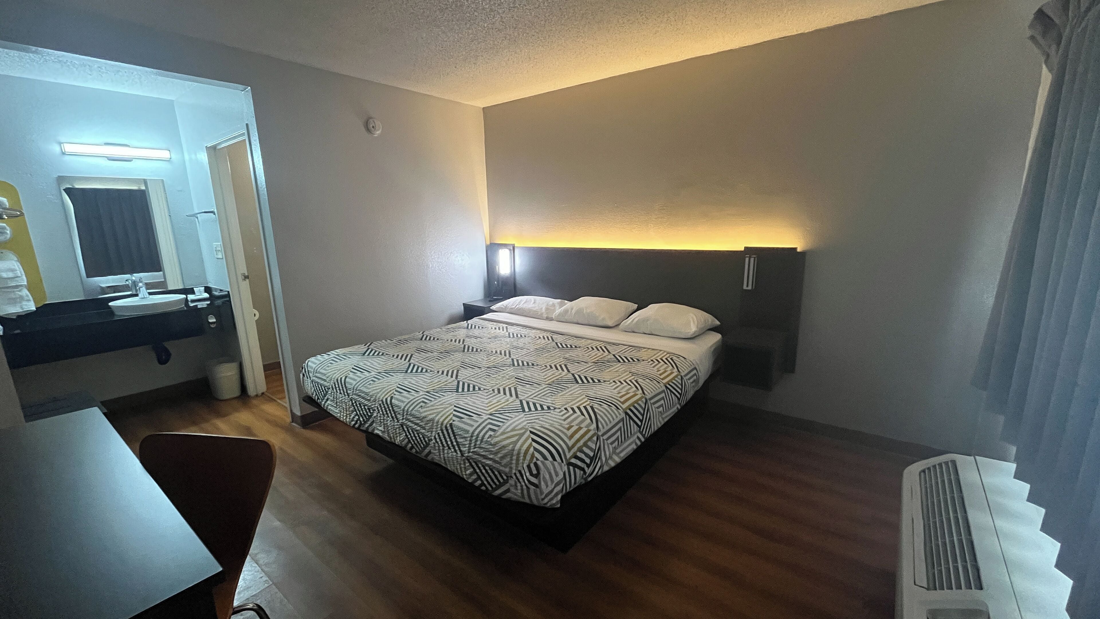 Una habitación de hotel con cama, mesita de noche y baño con lavamanos y espejo.