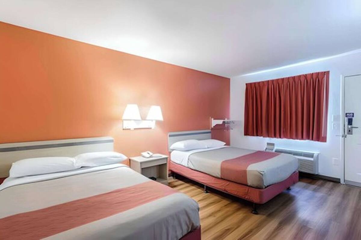 A hotel room with two beds, a nightstand, a lamp, a window with curtains, and a door.
