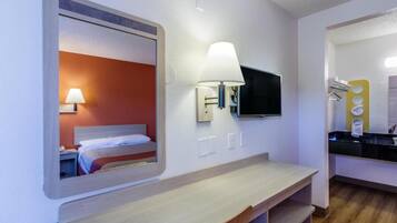A hotel room with a bed, a bedside lamp, a TV mounted on the wall, and a small kitchenette area.