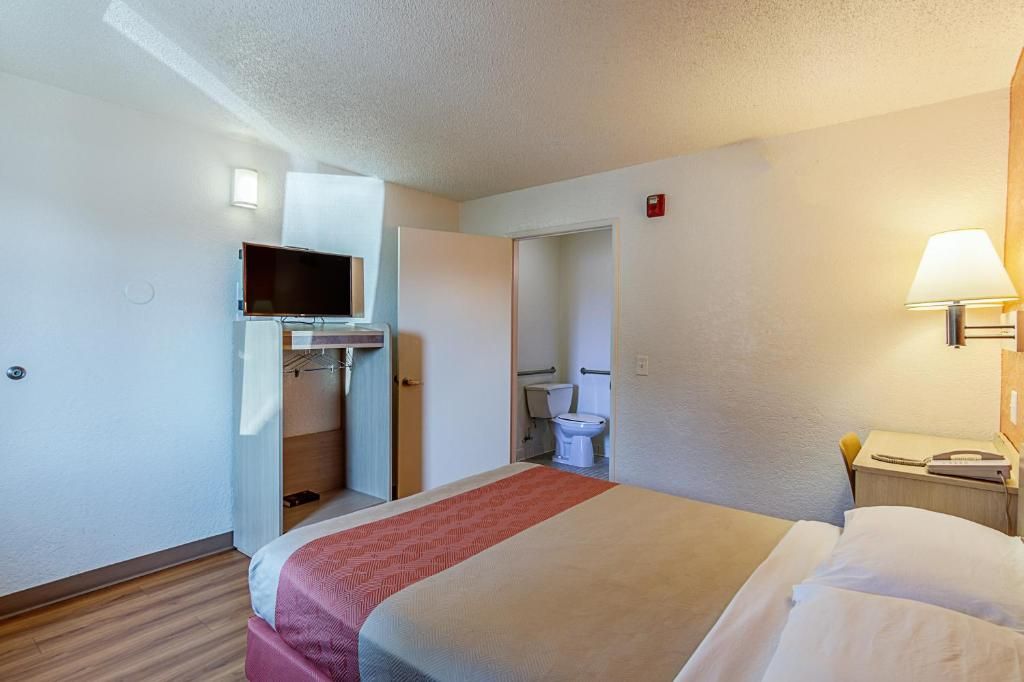 A hotel room with a bed, a television mounted on the wall, a bedside table with a lamp, and a bathroom visible through an open door.