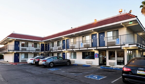 A multi-story motel with a red-tiled roof, a parking lot with several cars, and a blue handicap parking sign.