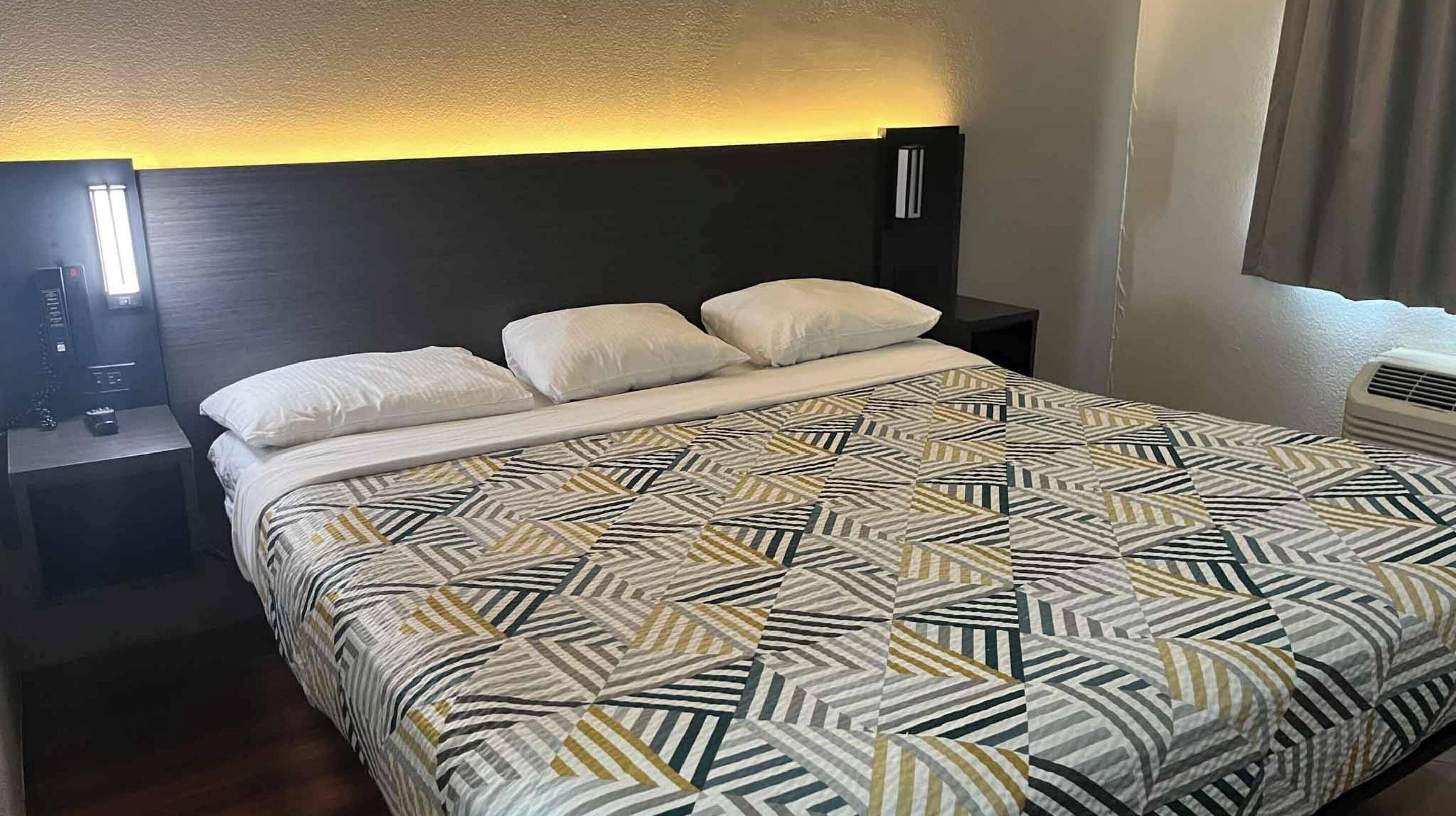 Quarto de hotel com cama, mesas de cabeceira, luminária fixada na parede e janela com cortinas.