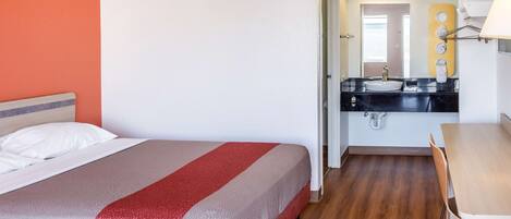 A hotel room with a bed, a desk, a bathroom with a sink and mirror, and a wooden floor.