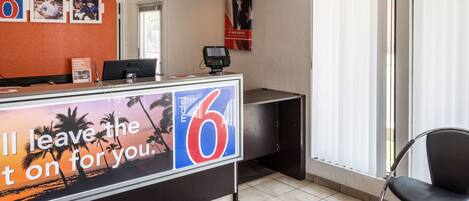 A hotel reception desk with a large '6' sign, a computer, and a phone. There are framed pictures on the wall and a window with blinds.