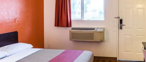 A hotel room with a bed, an air conditioning unit, a window with curtains, and a door.