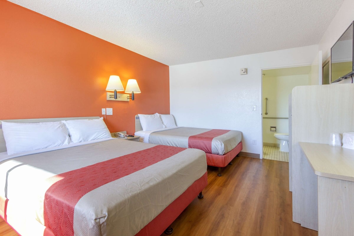 A hotel room with two beds, a wooden floor, a bathroom visible through an open door, and a small desk with a mirror.