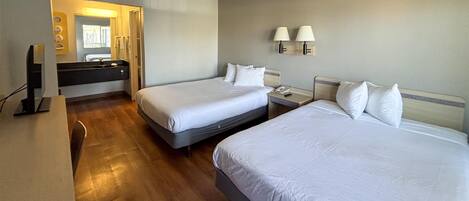 A hotel room with two beds, a desk, a mirror, and a bathroom with a sink and towel rack.