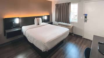 A hotel room with a large bed, bedside tables, a desk, a chair, a window with curtains, an air conditioning unit, and a door.