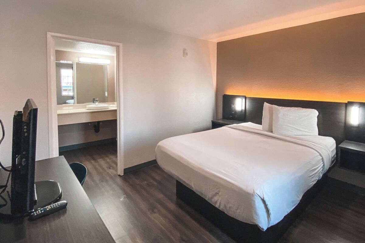 A hotel room with a bed, a desk with a TV and remote, a bathroom with a mirror and sink, and a nightstand with a lamp.