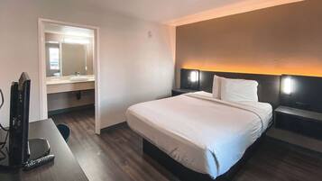 Habitación de hotel con cama, escritorio con televisor y control remoto, baño con espejo y lavamanos, y mesita de noche con lámpara.