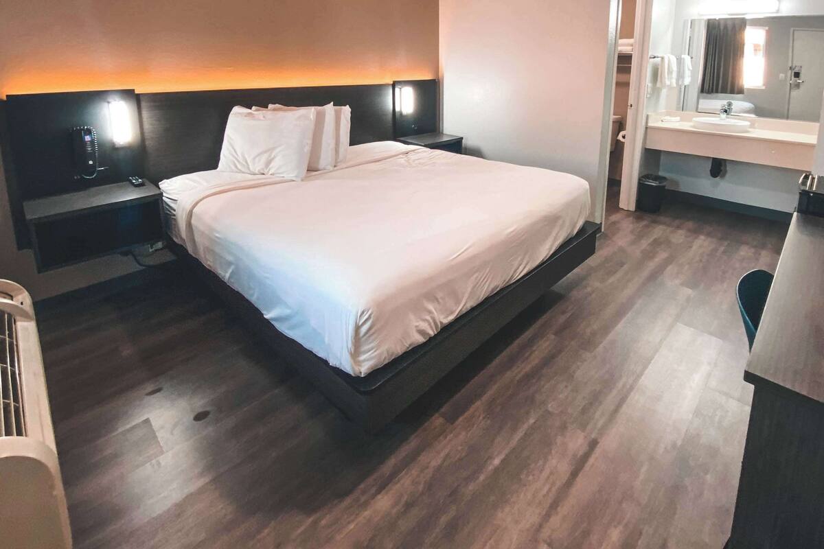 A modern hotel room with a large bed, bedside tables, a built-in desk, and a bathroom with a mirror and sink.