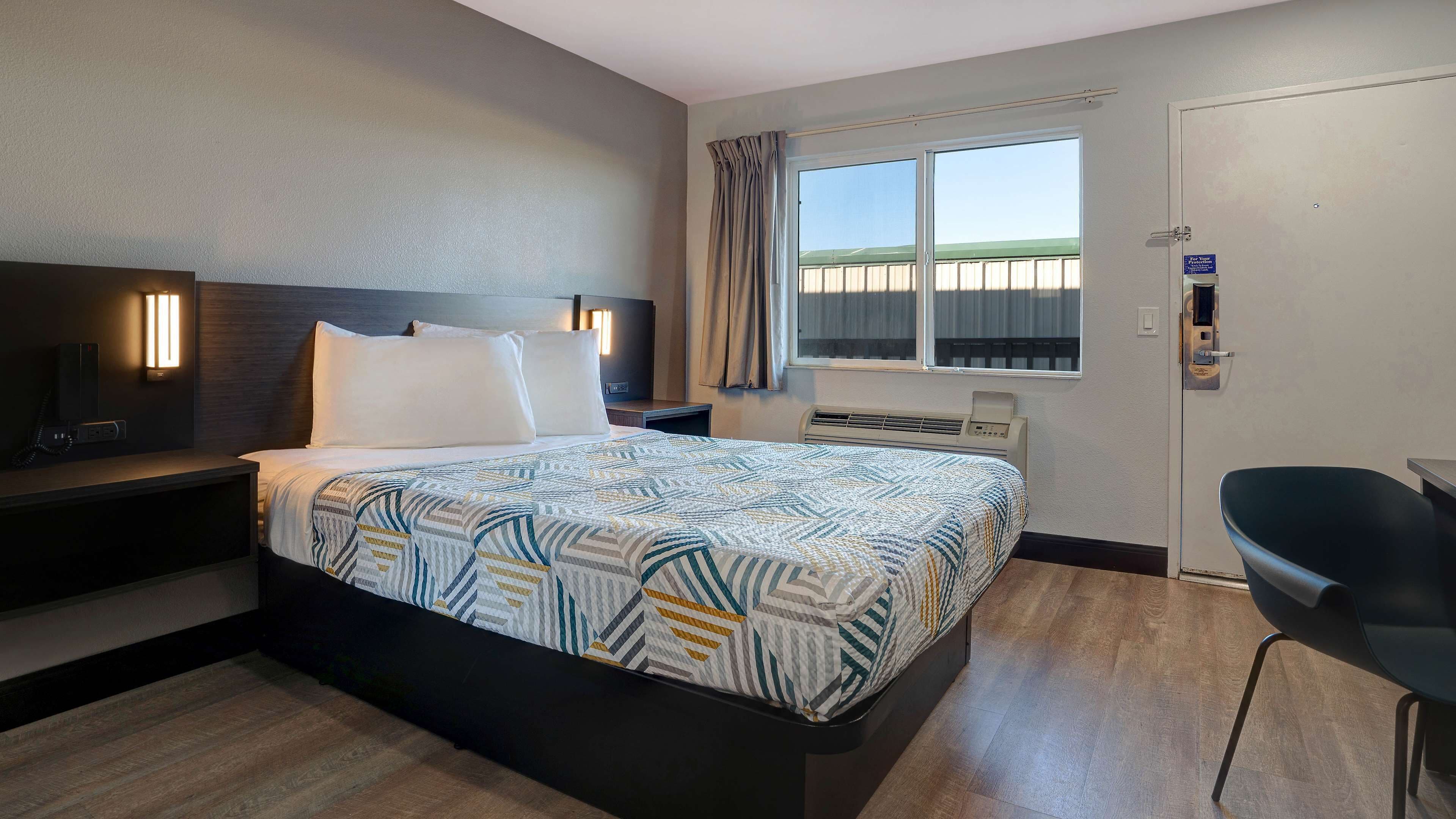 A hotel room with a bed, bedside table, lamp, air conditioning unit, window with curtains, and a chair.