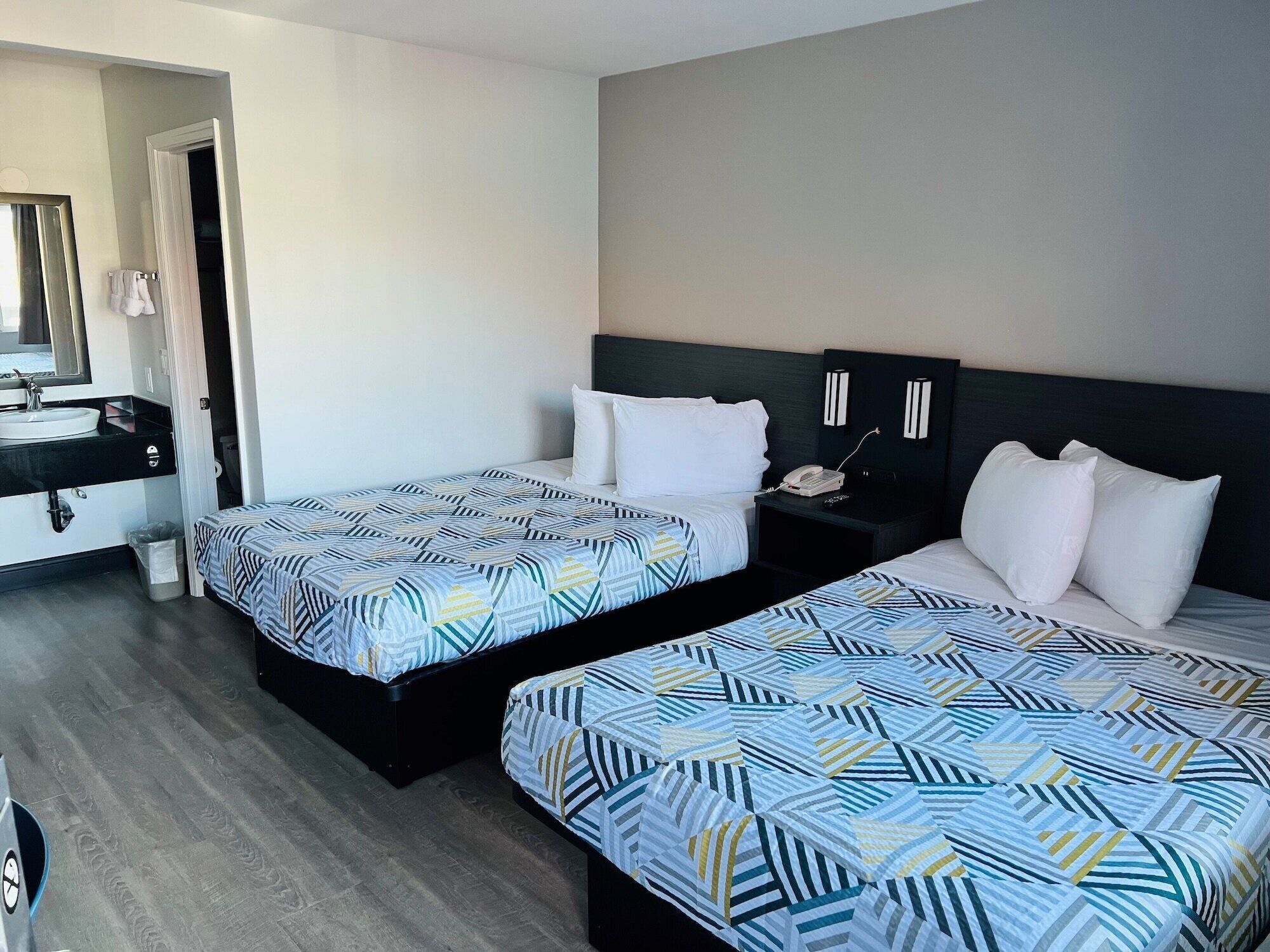 Habitación de hotel con dos camas, un baño con lavamanos y espejo, y una mesita de noche con teléfono.
