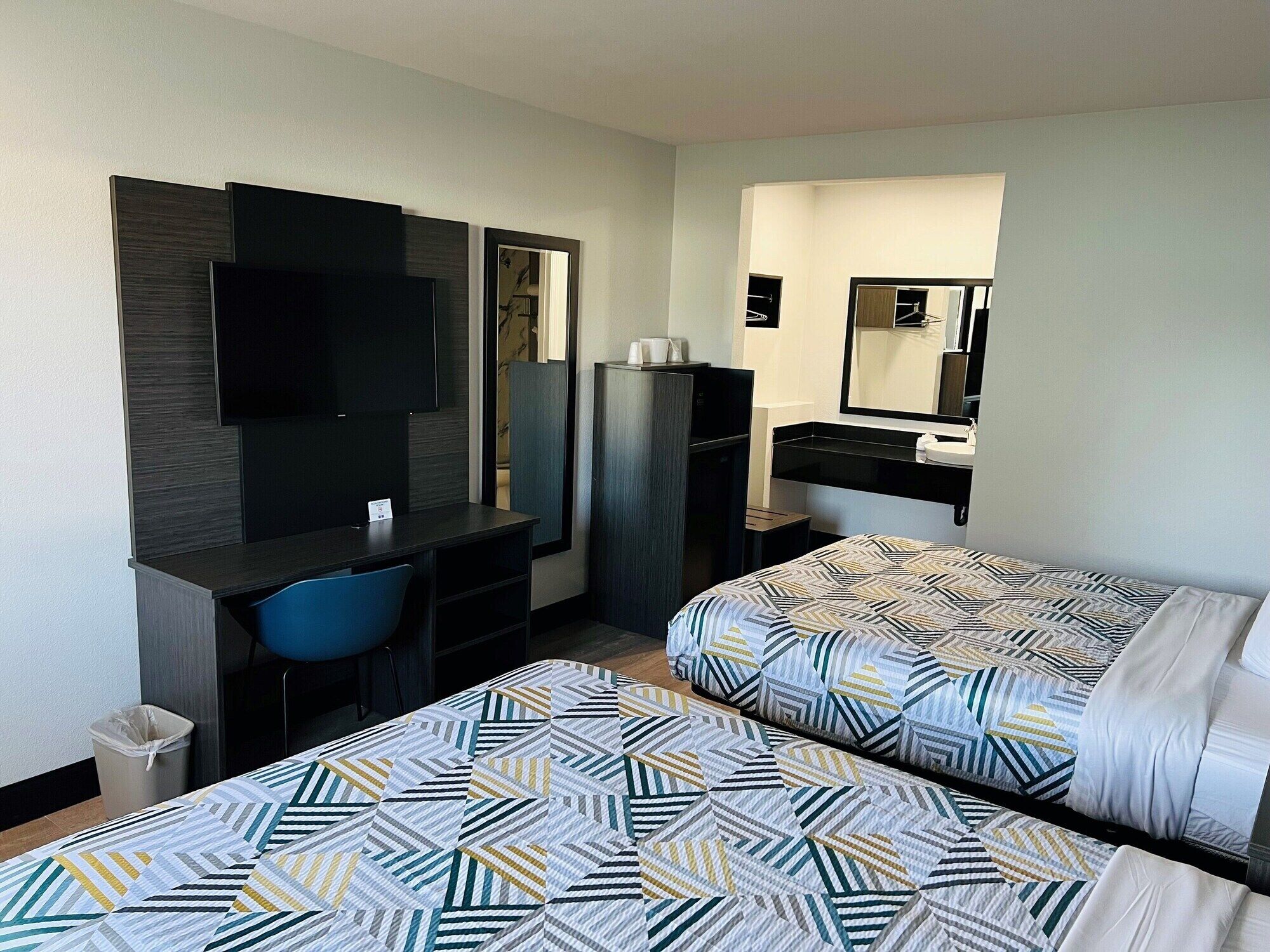 A hotel room with two beds, a desk, a chair, a TV, a mirror, and a small bathroom area.