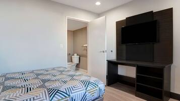 Una habitación de hotel con cama, televisor montado en la pared y baño visible a través de una puerta abierta.