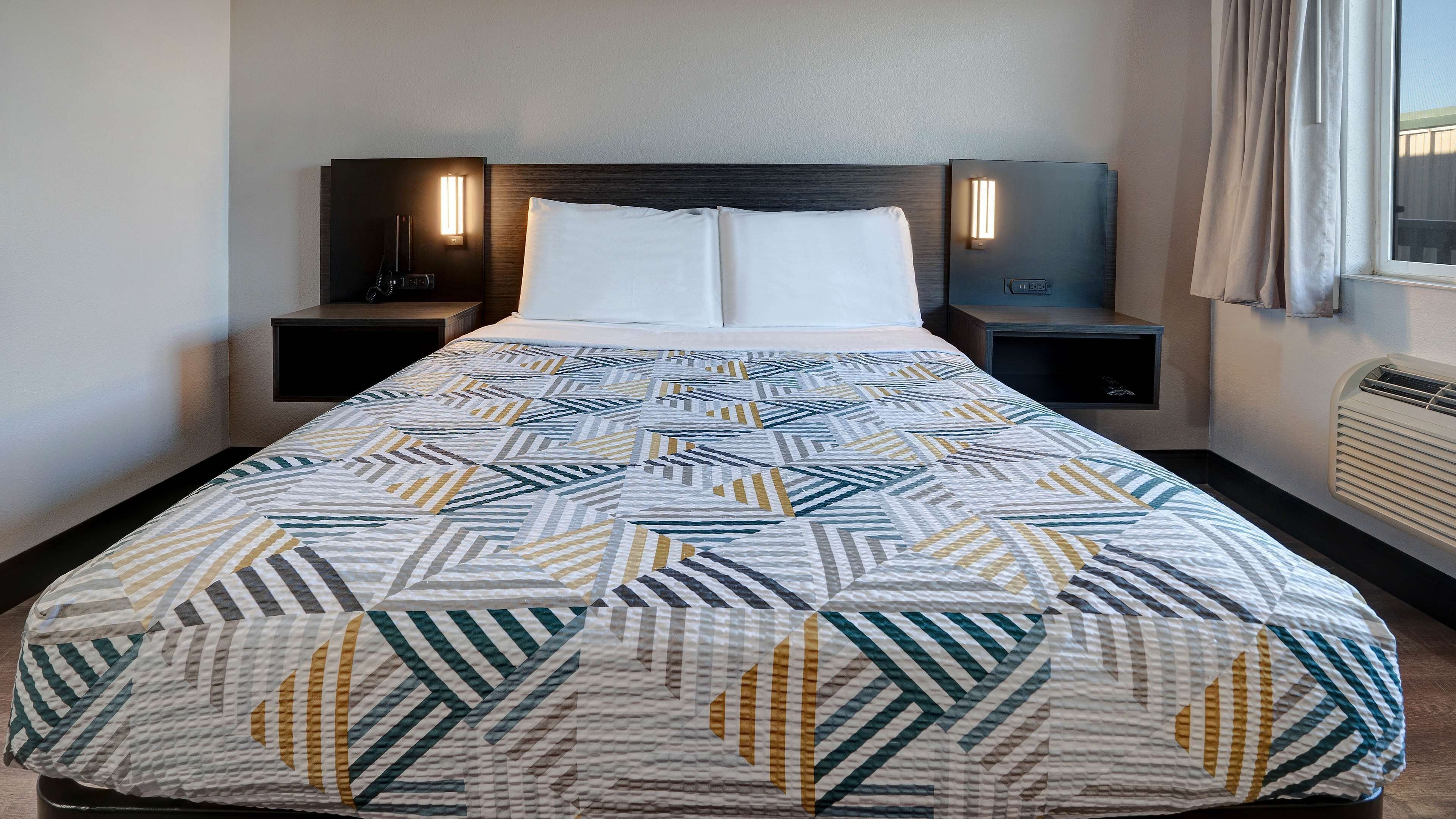 Uma cama bem arrumada com colcha estampada, duas mesas de cabeceira com abajures e uma janela com cortinas.