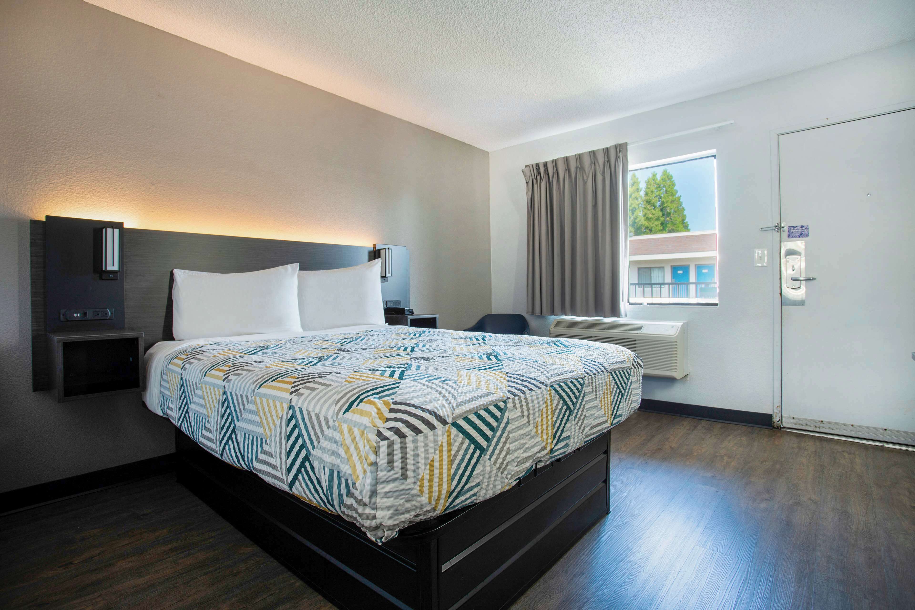 Una habitación de hotel con una cama, mesitas de noche, una ventana con cortinas y una puerta.
