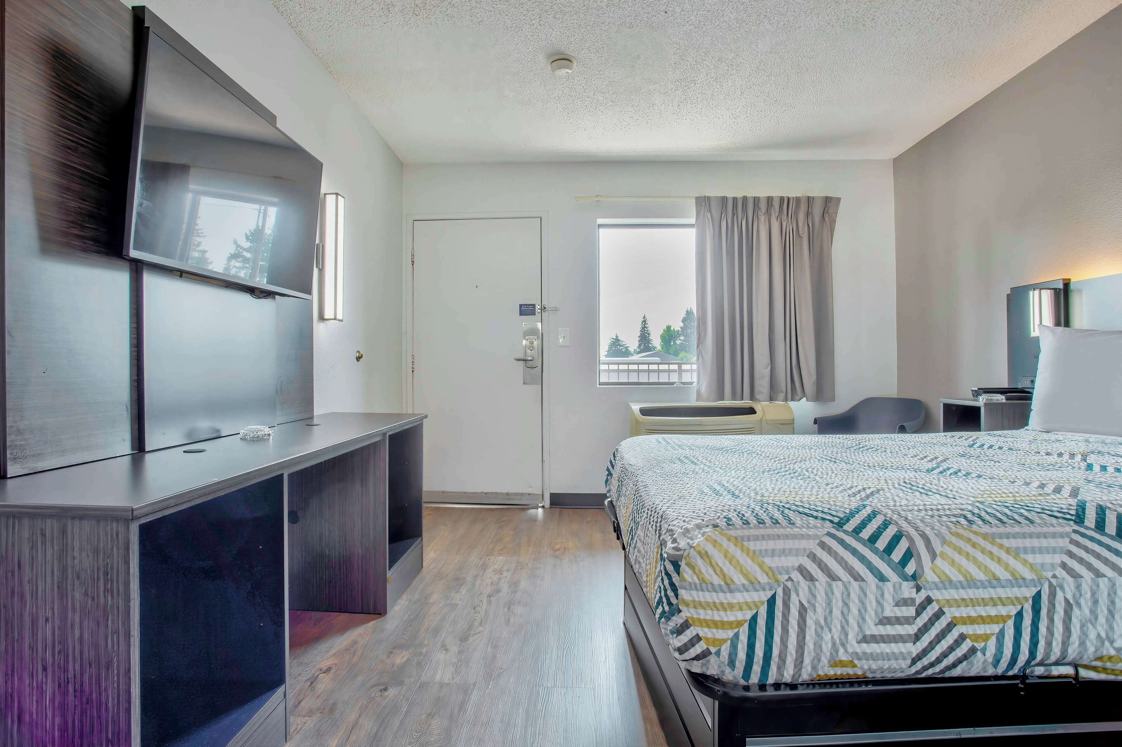 A hotel room with a bed, a desk, a TV, a window with curtains, and a view of trees.