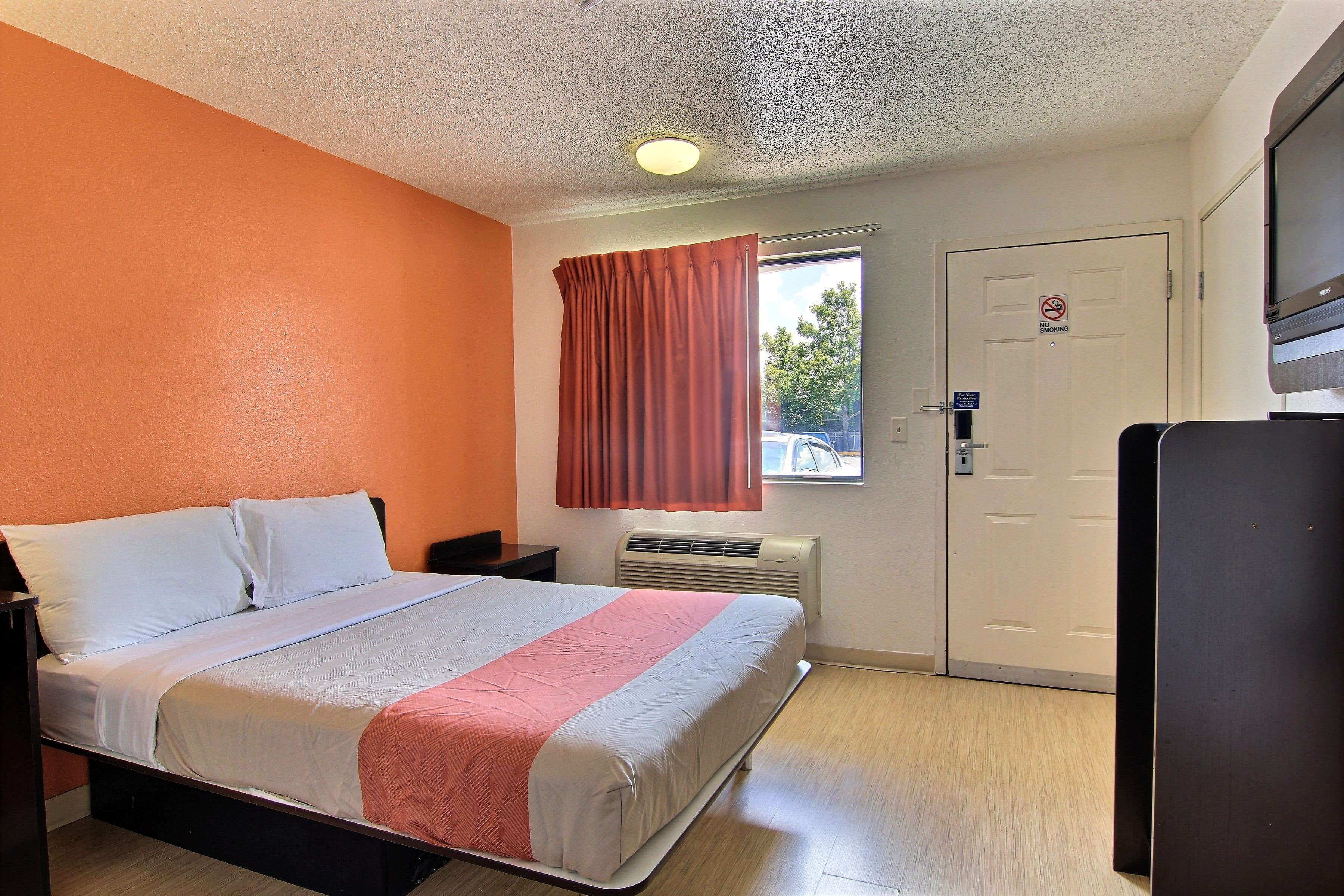 A hotel room with a bed, a window with curtains, a door with a 'No Smoking' sign, and a TV on a stand.