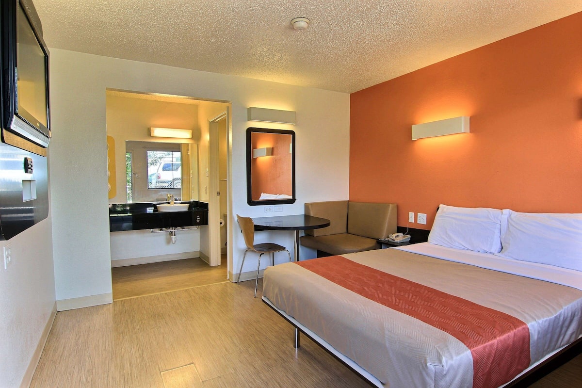 Quarto de hotel com cama, escrivaninha, cadeira, espelho e banheiro ao fundo.
