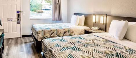 A hotel room with two beds, a window with curtains, a bedside table with a lamp, and a door with a sign.
