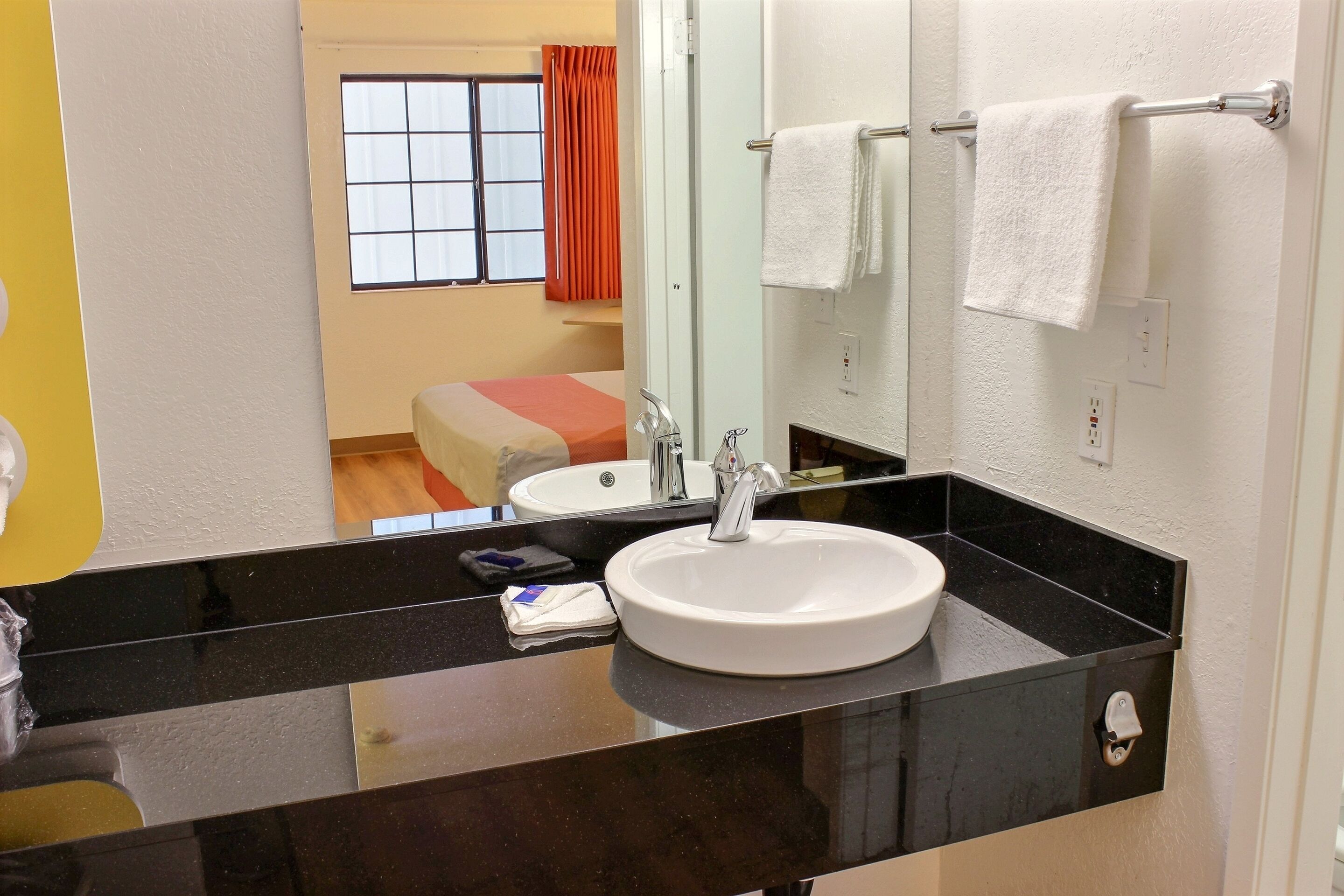 Une salle de bain avec un lavabo blanc, un plan de toilette noir et un miroir. On aperçoit une fenêtre avec des rideaux orange et, au fond, un lit recouvert d’une couverture à rayures rouges et blanches.