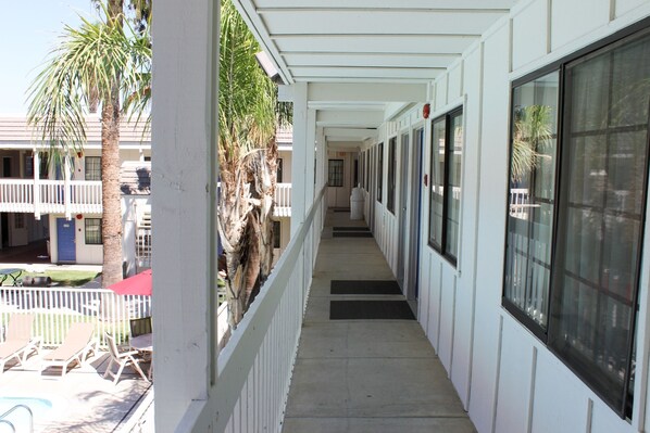 A corridor with a white railing, palm trees, and a pool area with lounge chairs. - Motel 6 Coalinga, CA - East (Coalinga)