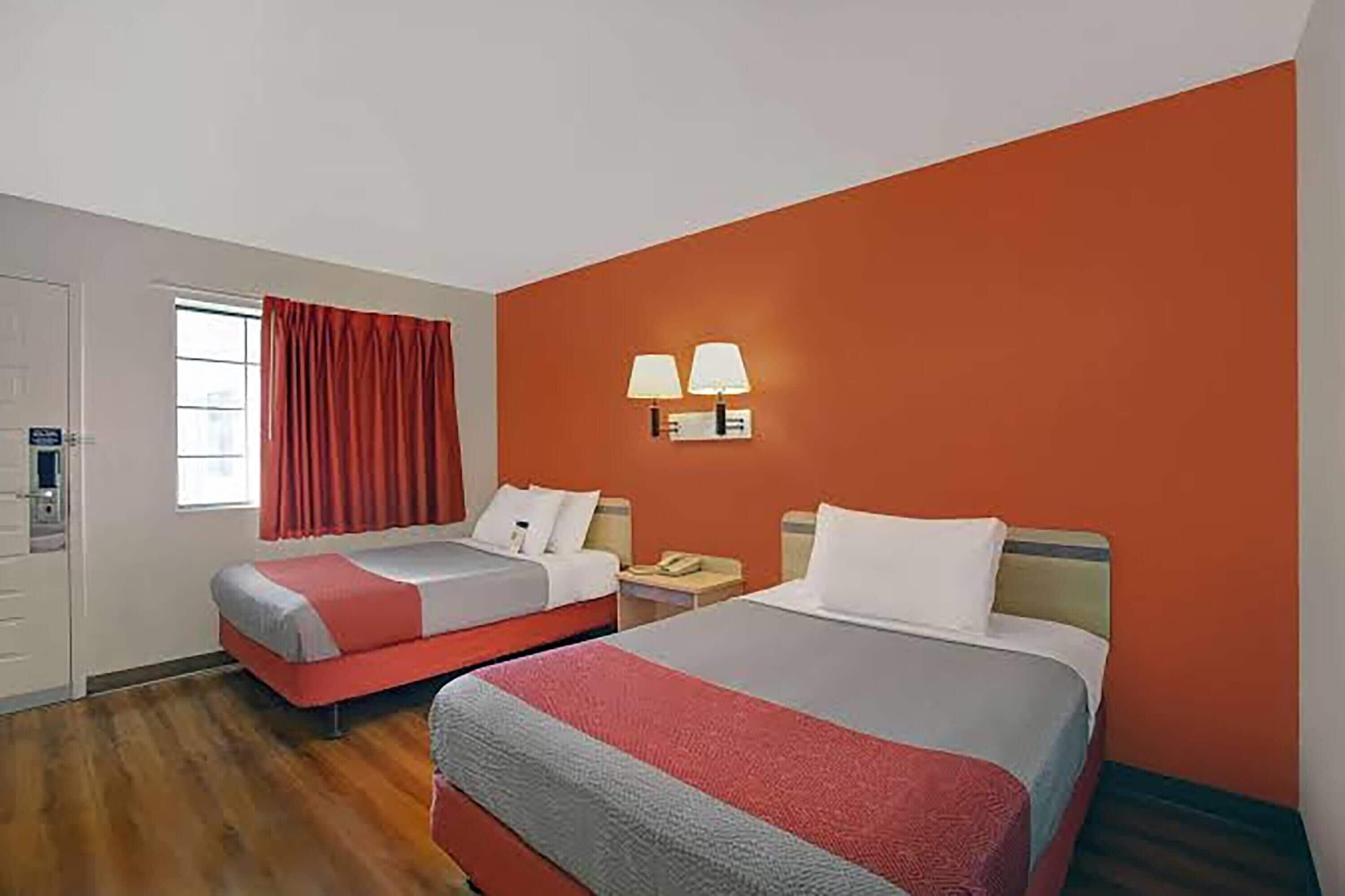 A hotel room with two beds, a window with red curtains, a wall-mounted lamp, and a wooden floor.