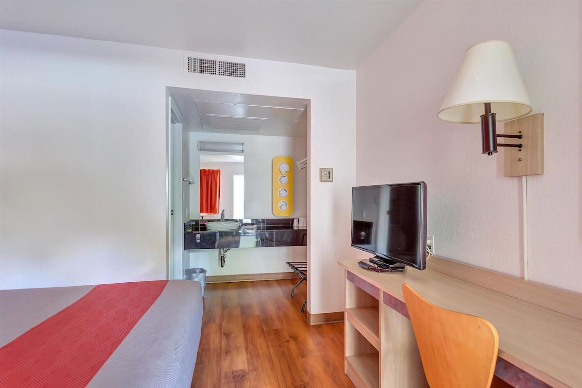 Une chambre d’hôtel avec un lit, un bureau et une chaise, une télévision, une lampe et une salle de bain visible à travers une porte ouverte.
