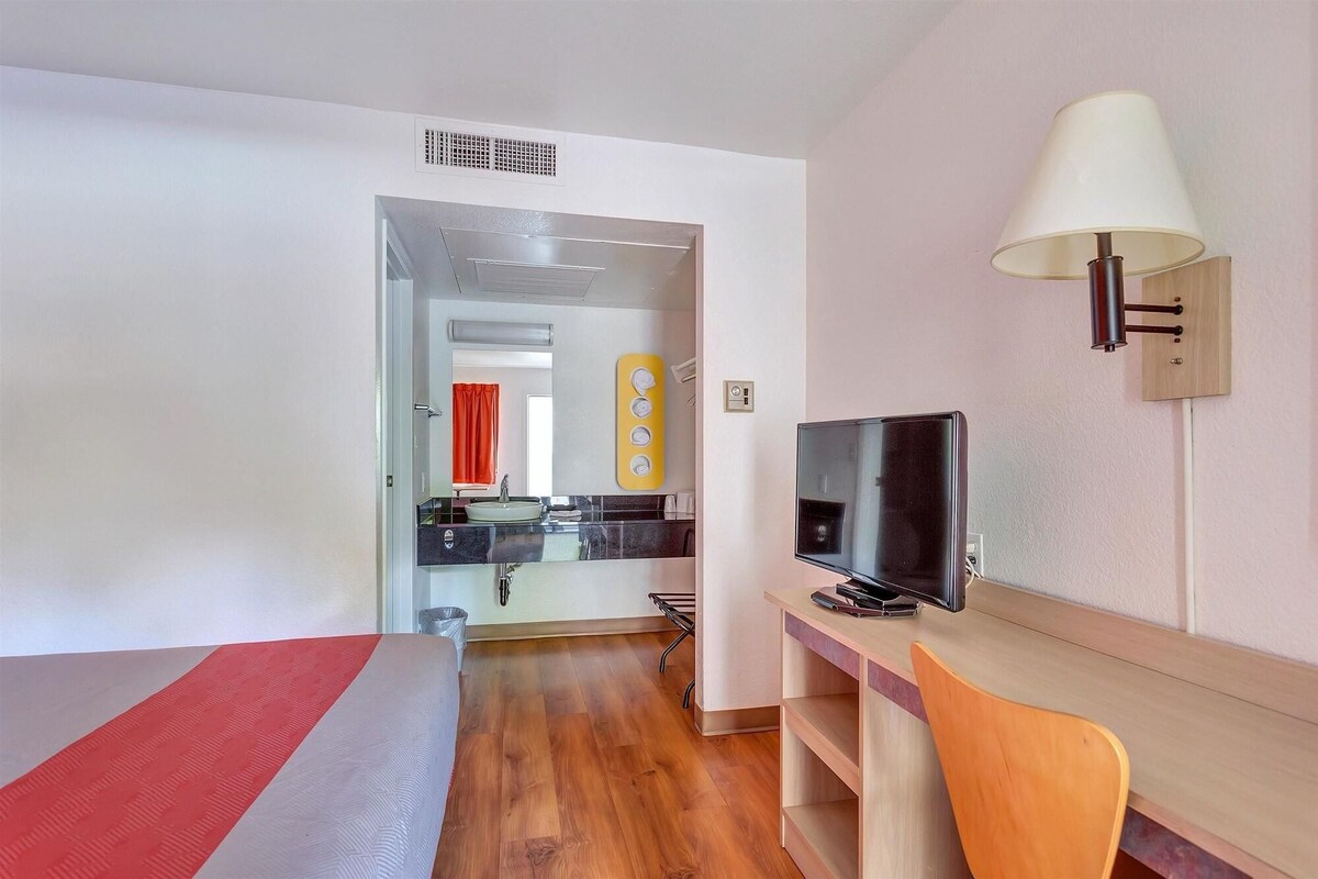 A hotel room with a bed, a desk with a chair, a TV, a lamp, and a bathroom visible through an open door.