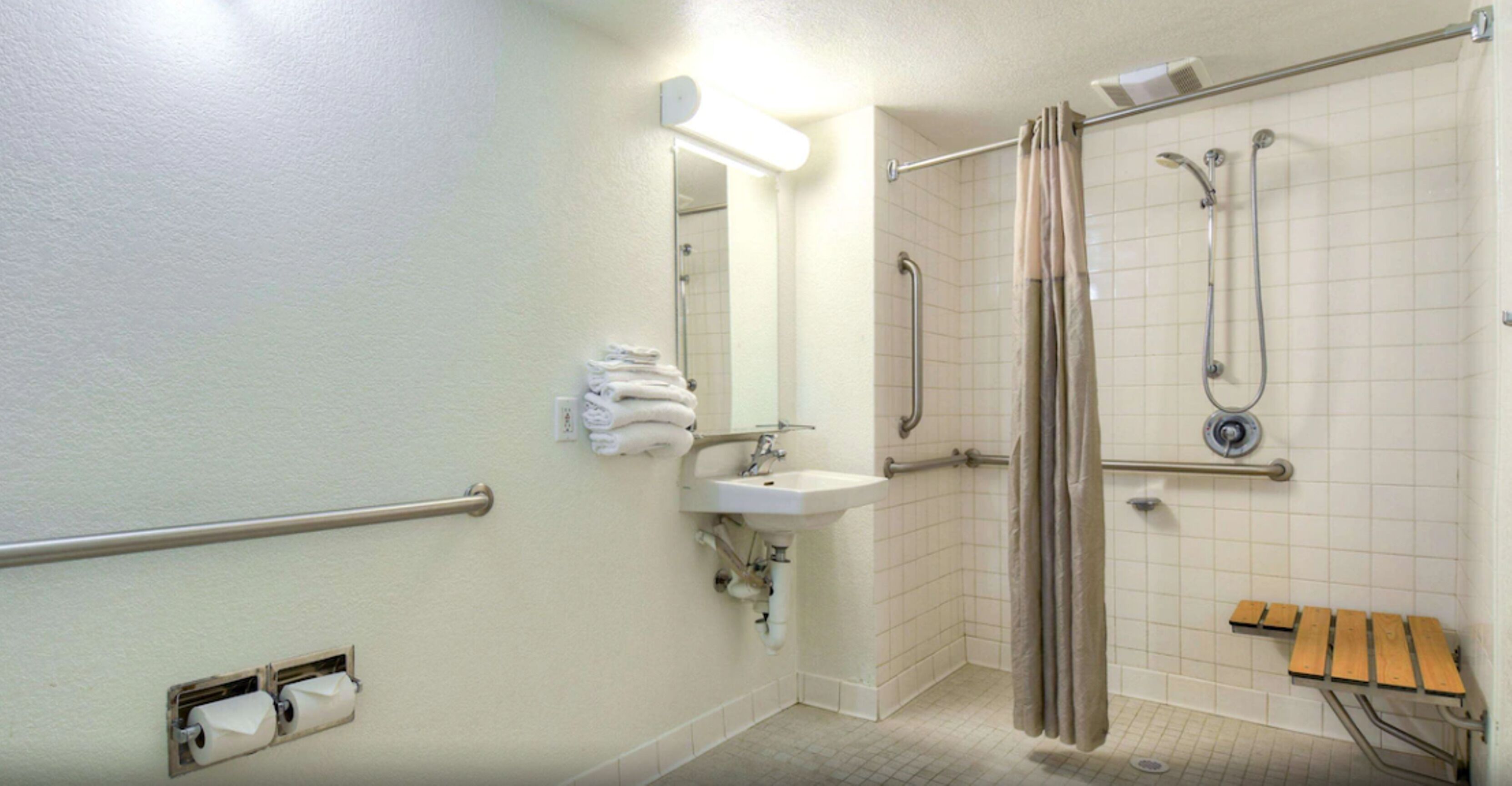 Un baño con ducha, barras de apoyo, lavamanos y dispensador de papel higiénico.